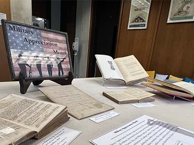 Selected books from the A.S. Salley Rare Book Collection lay opn in our first floor display case with a Military Appreciation Month sign printed with an American flag background.