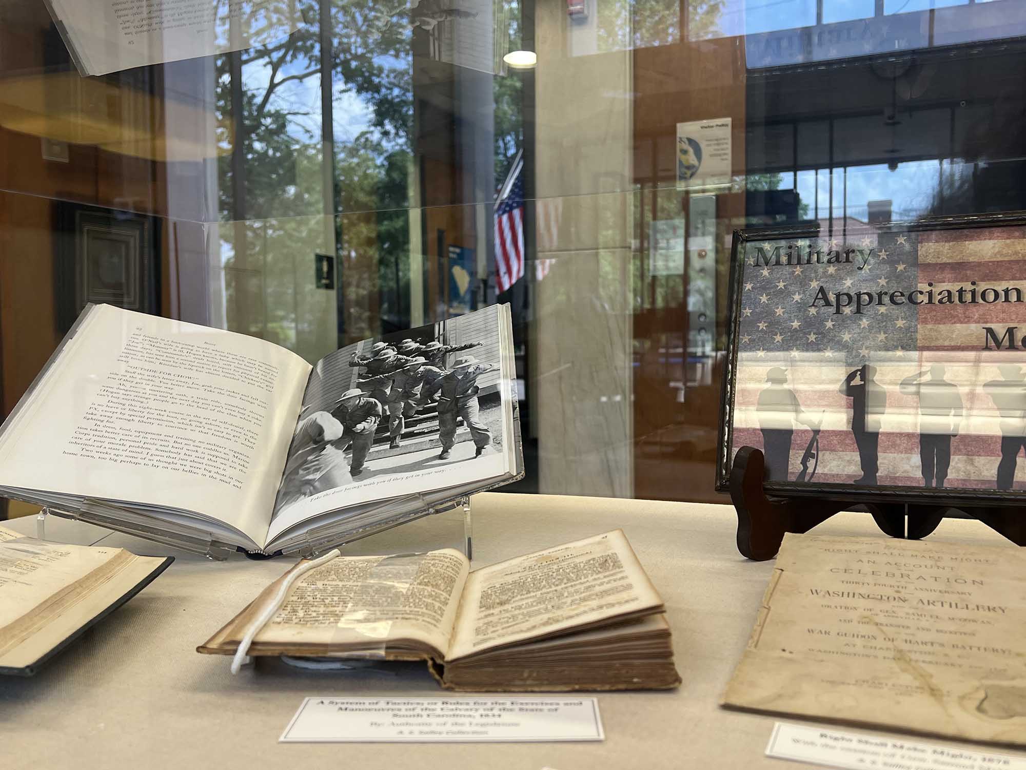 Selected books from the A.S. Salley Rare Book Collection are open in our glass display case with a framed Military Appreciation Month sign standing behind them.