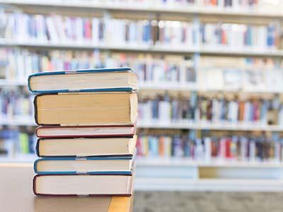 A stack of library books on a table, their spines not visible, and library shelves behind out of focus.