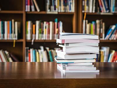 A stack of books on a table, library shelves visible behind them.