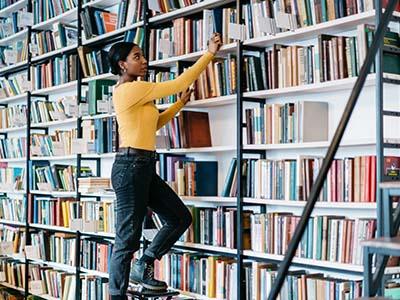 Dark skinned female librarian on a stool shelving books.