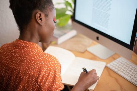 Woman writing in a notebook while working in front of a computer.