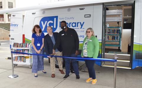 Wendy Coplen, YCL Director Jason Hyatt, van driver Brannon Nance and YCL Community Engagement Manager Dina Grant standing in front of the out reach van. They are about to cut a blue ribbon that is strung in front of the van. 